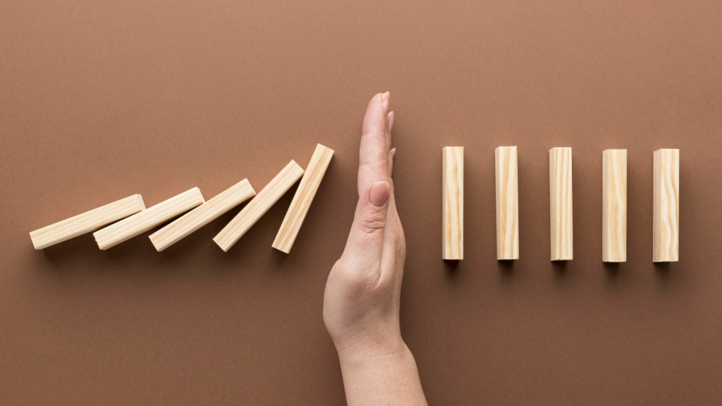 Une main arrête une série de blocs en bois désalignés sur un fond marron, symbolisant la prévention ou la maîtrise des déséquilibres.